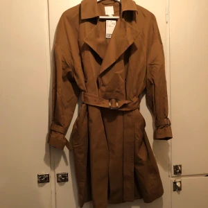 Grön/brun Trenchcoat från HM  - Grön/brun trenchcoat med skärp i midjan från HM. Aldrig använd (prislapp kvar). Nypris 599:-. Storlek XS men passar S. 
