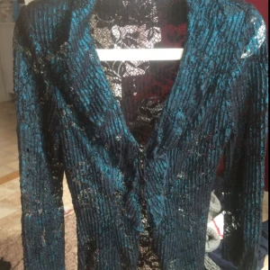  - En helt fantastisk vacker vintage kofta som är köpt i Berlin. Den är blå-skimmrande med glitter och transparent i ett fint mönster. Den har vadderade axlar och vida trekvarts-ärmar med en s.k "slit", dvs att ärmarna delar sig. Vid ärmarnas slut finns även ett snöre så kan man bestämma hur öppet man vill ha det där. (Se bild) Storleken är XS, funkar möjligen även om man är en liten S.  Tre knappar finns framtill att stänga koftan med (se bild). Koftan har även volanger längs med kragen och ner framtill.  