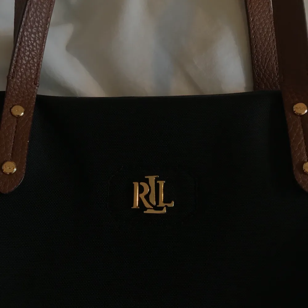En svart handväska från Ralph Lauren med bruna band och guldiga detaljer. Beige inuti med ett litet avtagbart fack för ex. telefon/kort. (Bild kan skickas vid intresse). Den är använd men fortfarande i bra skick. Mått ca: 44x28 cm⭐️. (Pris är exkl. frakt) . Väskor.
