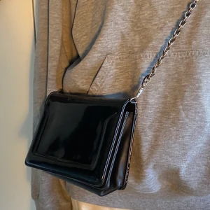 Svart axelväska - Liten axelväska i lack som passar till i princip allt. Plånbok och sådant får dock plats. Bandet som kommer med väskan är brytbart eftersom de knipsas fast i väskan. 