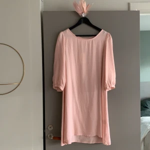 Klänning i rosa i fantastiskt material - Aldrig använd klänning i rosa. 100% i kupro (cellulosafabrik) som är lättskött, kan tvättas i maskin och skrynklar obetydligt. Fint skick. Ett band som man kan ha i midjan och ett spänne med fjädrar  etc följer med. 