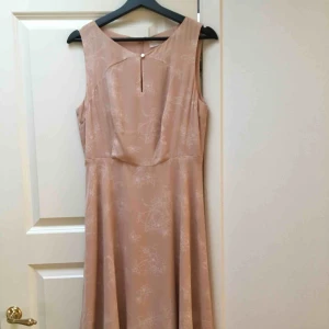  - Beige/sandrosa klänning från MQ i storlek 40. Använd sparsamt. Slutar vid knäna med en knapp vid bröstet. Dragkedja på ryggen. Blommigt mönster.