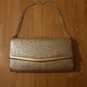 Guldig handväska  - Guldig handväska köpt på en loppis. Kom i en låda som verkar tillhöra. Lådan ger sken av att vara någorlunda gammal, men väskan har inga skador alls, verkar helt oanvänd. Säljer för 69kr exklusive frakt (44kr). Bjuder dock på frakten vid köp över 300kr. Skriv i dm vid intresse eller för frågor! 🥰