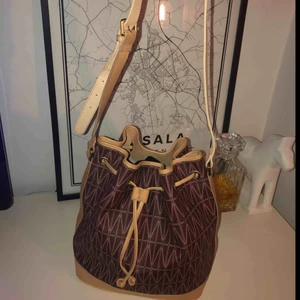  - Dagmars Bucket Bag är en klassisk och slitstark väska med Dagmars klassiska print i canvas. Väskan har tre innerfickor. Den har en justerbar axelrem i läder samt dragsko för stängning. Material: Canvas print med läder detaljer. Mått: 34cm x 30cm x 21,5cm.