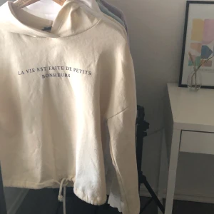 Hoodie - Beige hoodie från ginatricot med svart text. Fint skick, bara lite nopprig. Den har en snörning längs ned på tröjan. Köparen står för frakten.
