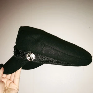  - Svart ”Emil-hatt” i filt med spännen på sidorna och flätade band över skärmen. Endast testad, ej använd.  Säljes pga fel storlek.  100kr + 30kr frakt. Betalning via Swish. 