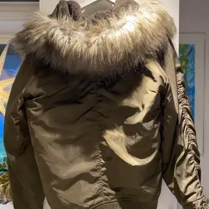 En tjockare militärgrön jacka från Pull&Bear med stor luva med fake päls. Två fickor framtill med en dragkedja och knappar, samt en liten kedja på vänster arm. Använd ett fåtal gånger 