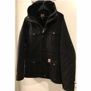 Carhartt Mentorjacket  - Supervarm höftlång jacka från Carhartt. Fodrad med svart fuskpäls i torso och i luvan. Dragsnöre i midjan på insidan om man vill ha en mer skuren form. Använd men mycket bra skick! Skickas mot porto el hämtas i Malmö. Obs, herrstorlek! 
