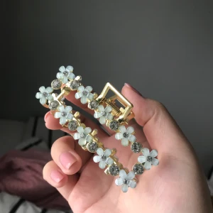 Ny guldig hårklämma med blommor och stenar - En metall klämma i guld färg. Jättesöt med stenar och ljusblåa blommor på sidan. Eftersom den är gjord i metall väger den lite mer än en vanlig hårklämma! Aldrig använd. Exklusive frakt