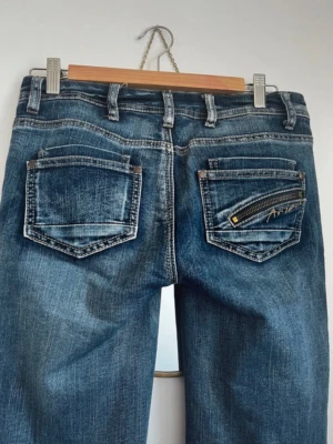 Blå Replay jeans med dragkedjedetalj - Säljer ett par blå jeans från Replay med snygg tvätt och raka ben. Bakfickan har en cool gulddragkedja och broderad Replay-logga. Klassisk femficksmodell med bälteshällor och kontrastsömmar. Perfekta för dig som gillar detaljer och streetstil.  Omsydda till lågmidjade, förra ägaren sa L men skulle säga M är bättre! Personen på bilden har Xs-S