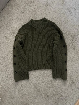 Olivgrön stickad tröja med knappar - Säljer en olivgrön stickad tröja med ribbad hög hals och snygga svarta knappar längs hela ärmarna. Tröjan har en croppad passform och är perfekt för lager på lager under höst och vinter. Tjockt garn och riktigt mysig vibe.