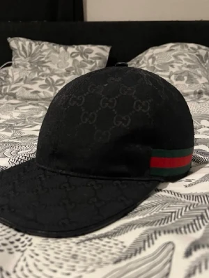 Svart Gucci keps med GG-mönster - Säljer en svart keps från Gucci med klassiskt GG-mönster över hela kepsen. På sidan finns den ikoniska gröna och röda randen. Justerbar rem bak i svart läder med guldfärgad spänne. Tillverkad i Italien och riktigt snygg detalj till din outfit.