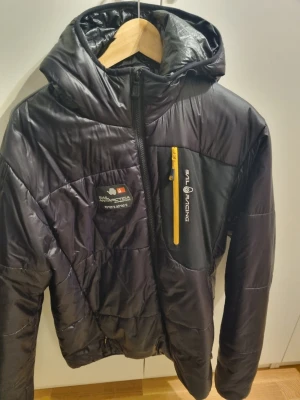 Svart pufferjacka från Sail Racing - Säljer en svart pufferjacka från Sail Racing med huva och gul dragkedja på bröstfickan. Fast pris.