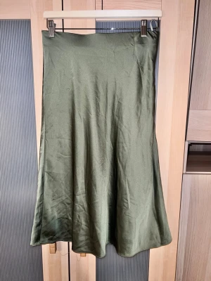 Olivgrön långkjol från Guess - Säljer en olivgrön långkjol från Guess i glansigt, mjukt material som ger en lyxig känsla. Kjolen har en enkel och stilren design utan mönster, med lätt utställd form som faller snyggt. Perfekt att styla med både sneakers och boots. Har använts några gånger därmed lösa trådar. 