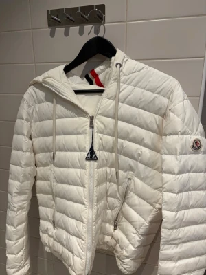 Vit Moncler pufferjacka med huva - Säljer en clean vit pufferjacka från Moncler med huva och coola detaljer som dragkedjor och snörning. Jackan har Moncler-logga på ärmen och snygga ränder i rött, blått och vitt på huvan. Perfekt för dig som vill ha en stilren och varm jacka. Storlek 2 i Monclers storlek så passar dig som har S/M