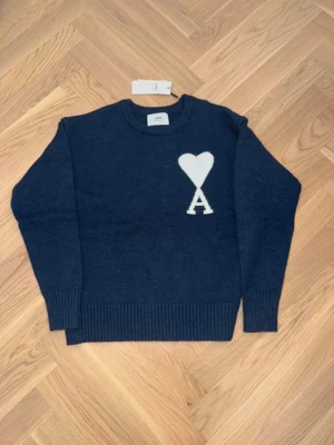 Mörkblå stickad tröja från Ami Paris - Säljer en mörkblå stickad tröja från Ami Paris Alexandre Mattiussi med ett vitt hjärta och bokstaven A på bröstet. Tröjan har rund hals, ribbade muddar och är långärmad. Perfekt för dig som vill ha en clean men ändå unik look.