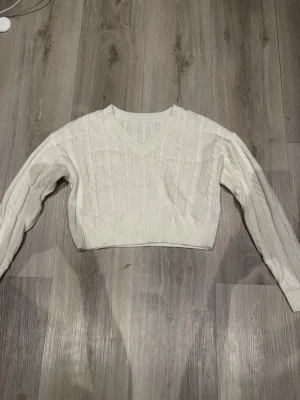 Vit croppad kabelstickad tröja - Säljer en vit croppad kabelstickad tröja med v-ringning och långa ärmar. Tröjan har ribbade muddar och ett klassiskt kabelmönster. Den är kort vid magen men med långa ärmar som sagt. Storlek xxs/xs 