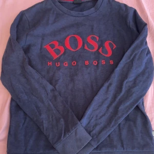 Blå långärmad tröja från Hugo Boss - Säljer en marinblå långärmad tröja från Hugo Boss med stort rött BOSS-tryck på bröstet. Tröjan har rund halsringning och ribbade muddar vid ärmslut och nederkant. Perfekt för dig som gillar stilrena logotröjor.