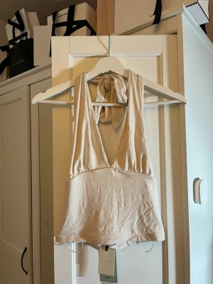 Beige omlott linnetopp med knyt - Supersnygg beige omlott linnetopp från Gina Tricot med djup v-ringning och breda axelband som knyts i nacken. Mjuk och stretchig känsla, perfekt till sommaren. Toppen är ärmlös och croppad för en trendig look.