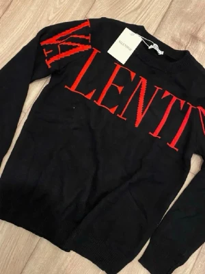 Svart stickad tröja från Valentino - Säljer en svart stickad tröja från Valentino med stora röda bokstäver över bröstet och axeln. Tröjan har rund halsringning och långa ärmar. Perfekt för dig som vill ha en statement piece i garderoben.