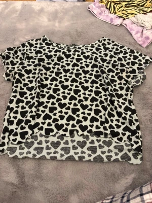 Vit topp med svarta hjärtan från Lindex - Säljer en söt vit topp från Lindex med svarta hjärtan över hela tyget. Toppen har korta ärmar och en lös passform. Materialet är mjukt och stretchigt, perfekt för dig som gillar lekfulla mönster. Nederkanten har en vågig finish som ger en extra detalj.