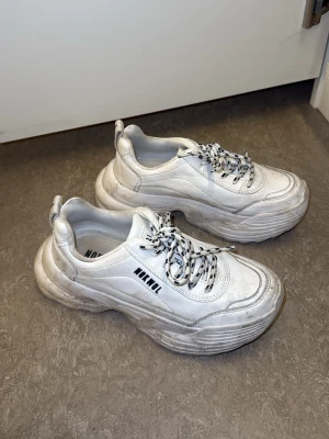 Chunky vita sneakers från NOKWOL - Säljer ett par chunky vita sneakers från NOKWOL med svartvitrandiga snören och tjock sula. Skorna har cool streetstyle-vibe. Dom behöver bara tvättas därav säljer billigt. 