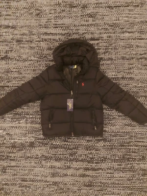 Svart pufferjacka från Polo Ralph Lauren (läs beskrivning) - Säljer en svart pufferjacka från Polo Ralph Lauren med huva och dragkedja framtill. Jackan har två fickor med dragkedja och den klassiska röda loggan på bröstet. Perfekt för kalla dagar och riktigt snygg till streetwear-stilen. (Skriv innan köp)