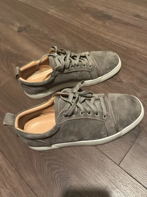 Grå mockasneakers från Christian Louboutin - Säljer ett par grå sneakers från Christian Louboutin i mocka med klassisk röd sula och vita kanter. Skorna har snörning, rund tå och beige innersida. Perfekta för dig som vill ha en clean och lyxig look med den ikoniska röda sulan.