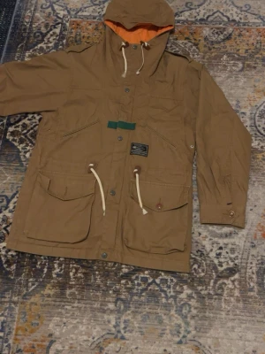 Brun parkajacka med huva och snörning - Säljer en brun parkajacka från General Patrol med orange innerfoder i huvan, stora fickor framtill och vita snören i midjan och huvan. Jackan har tryckknappar, dragkedja och detaljer i grönt samt bruna knappar på fickorna. Perfekt för dig som vill ha en chill och funktionell jacka.