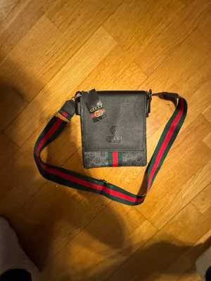 G väska  - Stilren crossbody-väska i mörk monogramcanvas med läderlock och klassiska gröna/röda detaljer. Perfekt storlek för vardagsbruk – rymmer mobil, plånbok och små essentials. Justerbar axelrem, kan bäras crossbody eller över axeln. Väskan är i bra skick och ger en clean, lyxig street-look som funkar till både vardag och outfit-fits. Säljs då den inte kommer till användning.