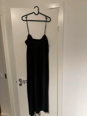 Svart långklänning från Gina Tricot - Säljer en svart långklänning från Gina Tricot med tunna axelband och snygg knytning framtill. Klänningen har en stilren och enkel design som ger en elegant look. Materialet känns mjukt och följsamt, perfekt för dig som gillar minimalistisk stil. Köptes från ginatricot, användes bara en gång. 