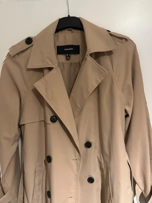 Beige trenchcoat från Vero Moda - Säljer en klassisk beige trenchcoat från Vero Moda med dubbelknäppning, stora slag och axelklaffar. Jackan har långa ärmar, knappar vid ärmslut och två fickor framtill. Perfekt för vår och höst när du vill ha en stilren look.
