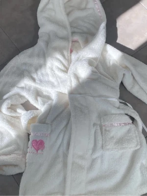 Vit mysig morgonrock/badrock med huva 💗 - Supermjuk vit morgonrock/badrock med huva och fina rosa detaljer samt broderade hjärtan. Perfekt efter dusch, bad eller för mys hemma. I fint skick, sparsamt använd. Har knytband i midjan och fickor framtill. ✨ Gullig & bekväm ✨ Perfekt som present ✨ Ren och redo att användas Storlek: one size  Skick: Mycket bra
