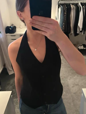 Svart kostymväst - Säljer en svart väst, öppen rygg. Västen har djup v-ringning och knäppning framtill. Sitter snyggt och tight, perfekt till jeans eller kjol. Storlek 38