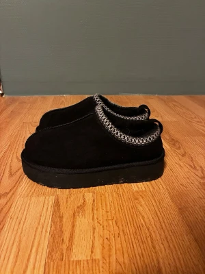 Svarta mockatofflor med grå kant - Mysiga svarta tofflor i mocka med tjock sula och dekorativ grå stickad kant runt öppningen. Perfekta för att hålla fötterna varma. Klassisk och enkel design med rund tå och platt sula.