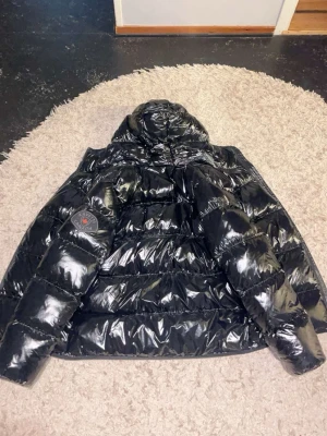 Svart glansig pufferjacka Zavetti Canada - Säljer en svart, glansig pufferjacka från Zavetti Canada med huva och patch på ärmen. Jackan har breda quiltade partier och dragkedja framtill. 