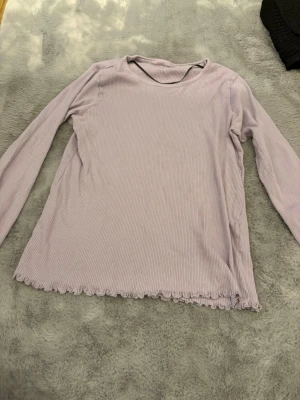 Ljuslila ribbad långärmad topp - Säljer en ljuslila ribbad långärmad topp med volangkant nertill. Toppen har en enkel rund halsringning och är tillverkad i ett mjukt, stretchigt material som sitter skönt på kroppen. Perfekt att styla med jeans eller kjol för en chill look.