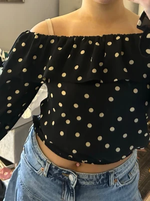 Svart offshoulder prickig blus - Säljer en svart offshoulder blus med vita prickar och volang upptill. Blusen är croppad och har långa, lätt transparenta ärmar med volang vid ärmslut. Perfekt för dig som gillar en lekfull och trendig stil.