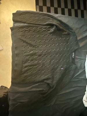 Svart kabelstickad tröja Polo Ralph Lauren - Kabelstickad svart tröja från Polo Ralph Lauren i storlek L. Klassisk rund halsringning och diskret broderad logga på bröstet. Tröjan har ribbade muddar vid ärmslut och nederkant, perfekt för dig som gillar stilrena och tidlösa plagg.