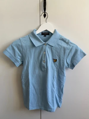 Ljusblå pikétröja från Lyle & Scott - Säljer en ljusblå pikétröja från Lyle & Scott med klassisk krage och tre knappar framtill. Tröjan har korta ärmar och den ikoniska gula loggan broderad på bröstet. Tillverkad i mjuk bomull som känns skön mot huden. Den är liten i storleken. Mer som en liten S. 
