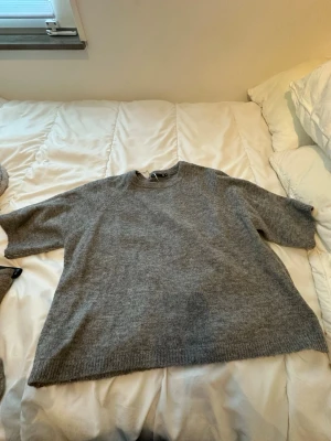 Grå stickad tröja från 157 - Mysig grå stickad tröja från 157 i storlek M. Tröjan har en enkel, rak passform med kortare ärmar och ribbade muddar vid ärmslut och nederkant. Perfekt för lager på lager under kyliga dagar.