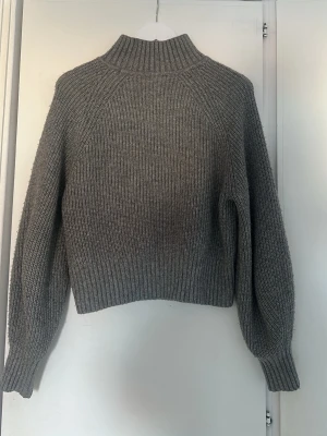 Grå ribbad stickad polotröja - Mysig grå stickad polotröja med ribbad struktur och hög krage. Tröjan har långärmade ärmar och en något boxig passform som ger en avslappnad vibe. Perfekt för kyliga dagar och enkel att styla med jeans eller kjol.