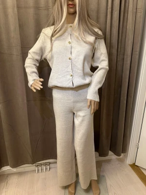 Beige stickad väst med guldknappar - Snygg beige stickad väst med rund hals och fyra guldiga knappar framtill. Västen har en enkel, rak passform och är gjord i mjukt material som känns skönt mot huden. Perfekt att styla över en t-shirt eller skjorta för en clean look.