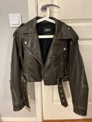 Croppad skinnjacka från Zara - Säljer en croppad skinnjacka från Zara i mörkgrön/brun-ish färg med coola detaljer som bälte i midjan, dragkedjor och stora slag. 