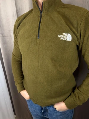 Olivgrön fleece från The North Face - Olivgrön fleece-tröja från The North Face med halv dragkedja och broderad logga på bröstet. Tröjan är långärmad och har en hög krage, perfekt för kyliga dagar. Mjuk och skön fleece som håller dig varm och bekväm.