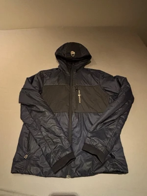 Svart-blå vindjacka från Sail Racing - Säljer en svart-blå vindjacka från Sail Racing i storlek medium. Jackan har huva, dragkedja framtill, två sidofickor med dragkedja och en bröstficka. Tillverkad i lätt polyester och perfekt för blåsiga dagar. Enkel och sportig design med logga på bröstet och huvan.