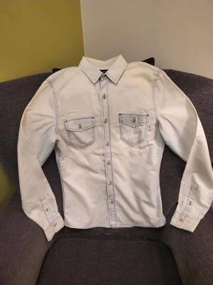 Vit jeansjacka från Bershka S - Säljer en vit jeansjacka från Bershka med kontrasterande svarta sömmar och snygga bruna knappar. Jackan har två bröstfickor med lock och knapp, klassisk krage och långa ärmar. Perfekt för dig som vill ha en clean men ändå edgy look.