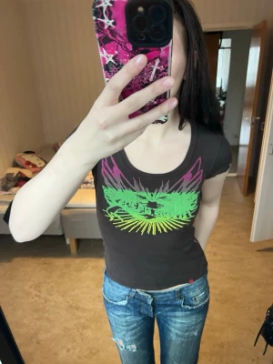 Brun t-shirt med neontryck från edc - Säljer en brun t-shirt från edc by Esprit med coolt neonfärgat tryck i grönt, gult och lila på bröstet. Modellen är figurnära med korta ärmar och rund halsringning. Perfekt för dig som gillar färgstarka detaljer och streetstyle.