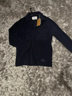 Mörkblå overshirt från NN07 - Snygg mörkblå overshirt från NN07. Helt nyköpt aldrig använt då lapparna sitter kvar. Tillverkad i mjuk ullmix som ger en clean look. Perfekt till våren och till dig som gillar stilrena detaljer.
