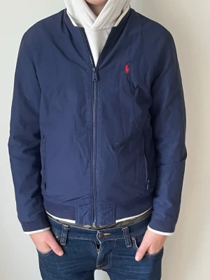 Marinblå bomberjacka från Polo Ralph Lauren - Säljer en stilren marinblå bomberjacka från Polo Ralph Lauren. Jackan har dragkedja framtill, två sidofickor och den klassiska röda loggan broderad på bröstet. Tillverkad i polyester och har en clean, sportig look som passar perfekt till jeans. Det är storlek l i barnstorlek
